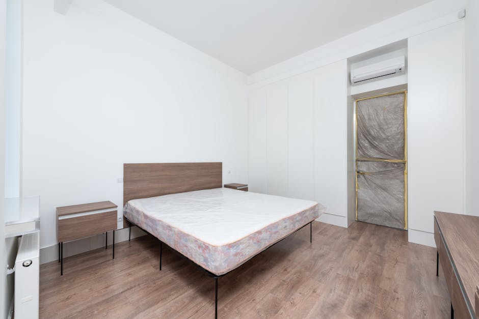 A minimalist bedroom featuring a double bed with a wooden headboard and a plain mattress, positioned centrally against a white wall. On either side of the bed are small matching wooden nightstands with simple, flat surfaces. The room has wooden flooring with a smooth finish, and there is a white wardrobe built into the wall, with tall, sleek panels. A window is covered with a protective plastic sheet framed in gold tape, indicating ongoing renovation or preparation for cleaning or renovation. An air conditioning unit is mounted above the window, contributing to the room's climate control. Soft, natural lighting illuminates the space, highlighting the clean, tidy condition of the surfaces and furnishings. The overall scene emphasizes a well-maintained, hygienic environment suitable for professional domestic cleaning, serviced by Carpet Cleaning Pimlico, as part of their surface cleaning and sanitisation processes.