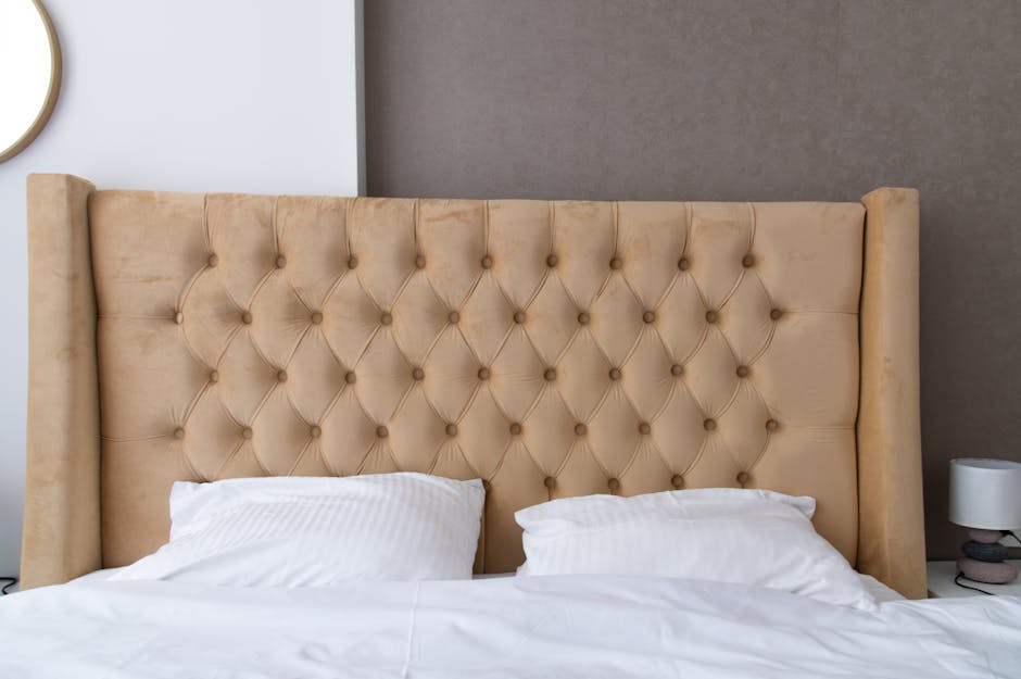 Close-up of a neatly made bed in a bedroom, featuring a tall, beige, tufted upholstered headboard with diamond-patterned buttons. The bed is covered with white linens, including two pillows with striped pillowcases. On the right side of the image, there is a small white bedside lamp placed on a dark-colored nightstand. The wall behind the headboard is painted in a neutral taupe or light brown shade, and the room is softly lit, emphasizing the clean and tidy condition of the surfaces. Carpet Cleaning Pimlico specializes in domestic surface cleaning, ensuring hygienic and well-maintained bedroom environments.