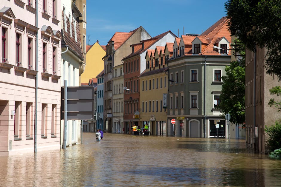 A group of emergency responders and workers in protective clothing are gathered in a residential street during a flood response, with large sandbags stacked against a wall to prevent further water intrusion. A silver car is parked on the wet asphalt, which reflects the surrounding houses, utility poles, and trees. The scene is set under an overcast sky with diffuse lighting, and neighboring houses with tiled roofs and trees are visible in the background. The area appears damp and involved in flood mitigation efforts, highlighting the importance of surface cleaning and disaster response by [COMPANY_NAME].