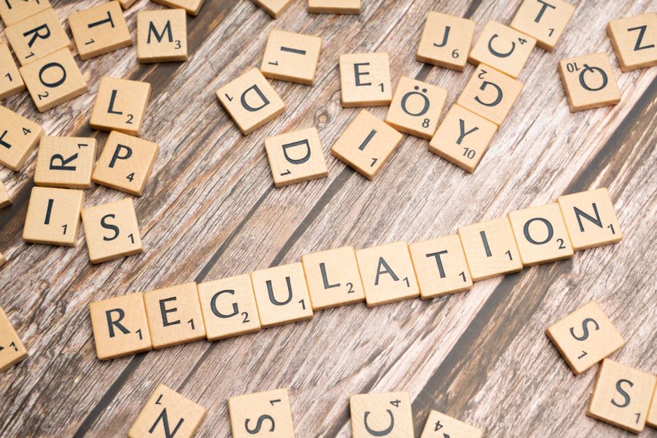 A close-up of a wooden surface with scattered Scrabble tiles, some face down and others showing letters. The tiles spell out the word 'REGULATION' in the center, with additional tiles surrounding it. The surface appears to be a rustic wooden table with visible grain and texture, illuminated by soft, natural lighting, emphasizing the clean and organized appearance typical of professional cleaning and hygiene standards. The image relates to the importance of adhering to legal requirements in commercial cleaning, as highlighted on the Carpet Cleaning Pimlico website, which specializes in cleaning services in the Pimlico area.
