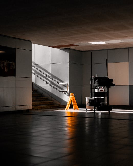 The image depicts an indoor commercial space with a tiled floor made of dark, polished ceramic reflecting light and shadows. In the background, a staircase with wooden steps and stainless steel handrails ascends along a wall composed of large, smooth, light grey panels. To the right of the staircase, there is a cleaning trolley equipped with various cleaning supplies, including spray bottles and cloths, situated in front of a sectioned wall with a beige panel. An orange wet floor caution sign is prominently placed on the floor near the trolley, indicating recent or ongoing cleaning activities. The overall scene is illuminated by ceiling lighting, creating a clean, professional environment associated with surface cleaning and maintenance, as emphasized by the presence of the cleaning equipment. Carpet Cleaning Pimlico offers expert deep cleaning and sanitisation services for commercial spaces such as this, ensuring hygiene and surface longevity.