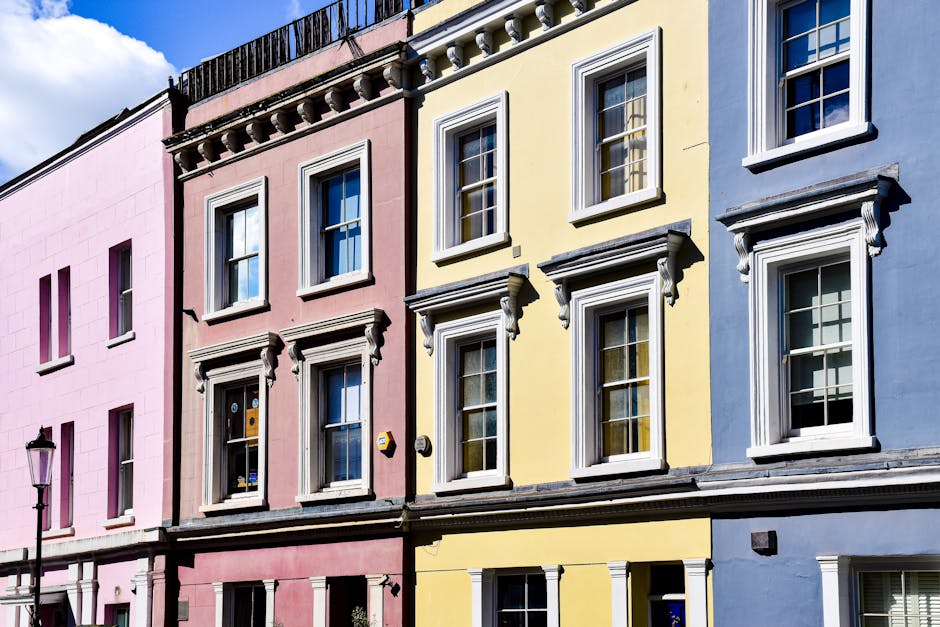 An exterior view of a row of three terraced houses featuring pastel-colored facades in pink, yellow, and blue. Each house has white-framed sash windows with decorative molding and shutters, and the buildings are illuminated by natural daylight against a partly cloudy sky. The image showcases the clean, well-maintained surfaces of the buildings, emphasizing the smooth painted exteriors and tidy window panes, with a vintage streetlamp visible on the left side. While the focus is on residential architecture, [COMPANY_NAME] specializes in surface cleaning and deep cleaning services, including sanitisation, for similar homes in Pimlico, ensuring their facades and interiors maintain their appearance and hygiene.