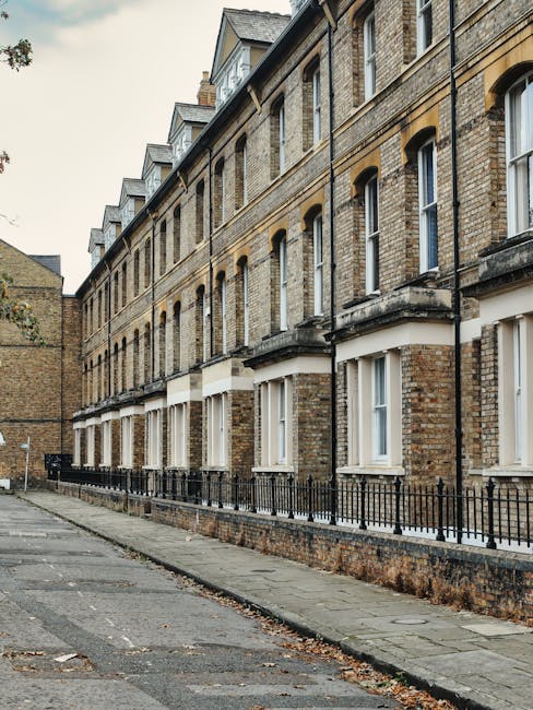Exterior view of a row of traditional brick terraced houses in Pimlico, featuring large sash windows with white frames and black wrought iron railings along the sidewalk. The pavement appears clean with minimal debris, and the buildings exhibit a weathered appearance with neatly maintained facades. The scene is illuminated by natural daylight, highlighting the textured brickwork and architectural details. This image reflects a typical residential street where professional domestic cleaning, including surface cleaning and maintenance of the building exteriors, is essential to uphold hygiene and aesthetic appeal, as offered by Carpet Cleaning Pimlico.