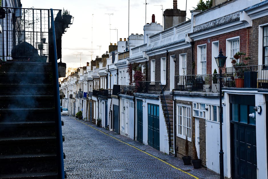 An exterior view of a row of three terraced houses featuring pastel-colored facades in pink, yellow, and blue. Each house has white-framed sash windows with decorative molding and shutters, and the buildings are illuminated by natural daylight against a partly cloudy sky. The image showcases the clean, well-maintained surfaces of the buildings, emphasizing the smooth painted exteriors and tidy window panes, with a vintage streetlamp visible on the left side. While the focus is on residential architecture, [COMPANY_NAME] specializes in surface cleaning and deep cleaning services, including sanitisation, for similar homes in Pimlico, ensuring their facades and interiors maintain their appearance and hygiene.