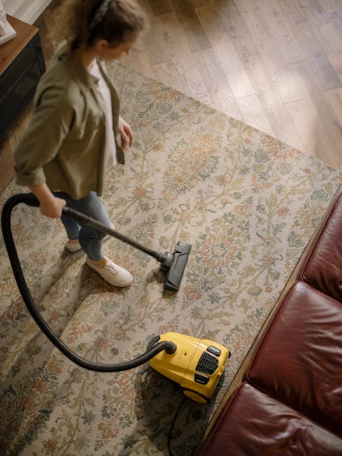 A professional cleaning specialist from Carpet Cleaning Pimlico is performing deep surface cleaning on a beige, plush carpet in a bright, well-lit living room. The worker is dressed in a full white protective suit, gloves, a face mask, and boot covers, using a large vacuum or extraction machine with a hose to thoroughly clean the carpet. The room features a white wall with a window allowing natural light, a wooden shelving unit with decorative items, and potted indoor plants, all giving a tidy and hygienic appearance. The scene emphasizes domestic cleaning and sanitisation processes aimed at maintaining a clean, fresh environment near SW1V, Pimlico.