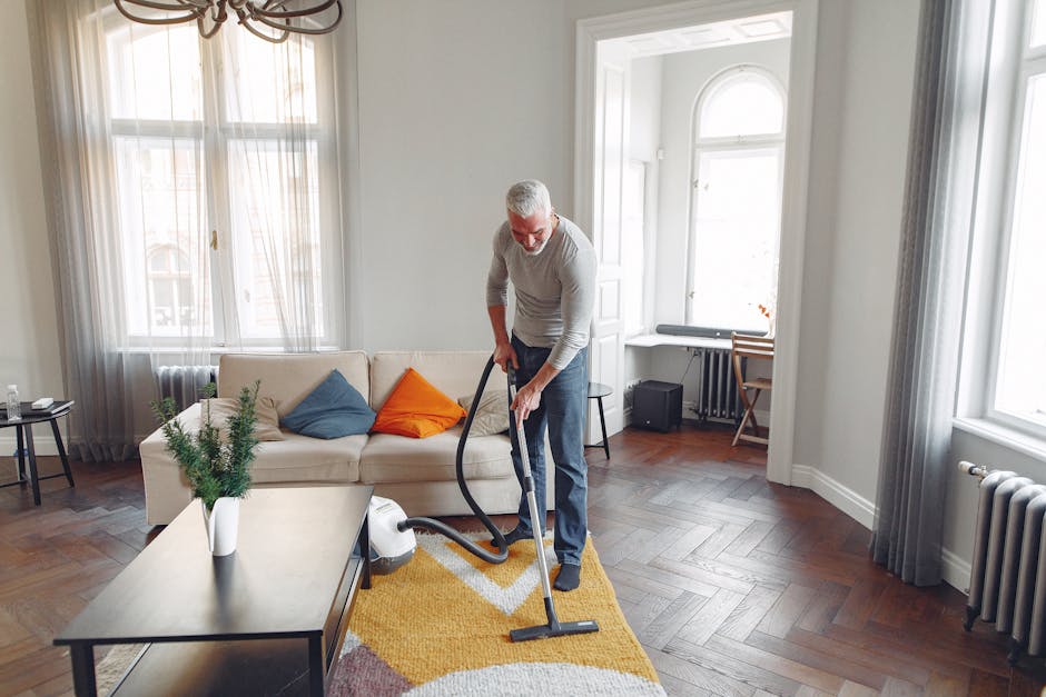 A man performing surface cleaning in a bright, spacious living room with large windows allowing natural light to fill the space. The room features a white sofa adorned with colorful cushions, a wooden coffee table with a potted plant, and hardwood flooring with a yellow and white patterned rug. He is using a vacuum cleaner, part of a professional cleaning process by Carpet Cleaning Pimlico, to thoroughly clean the wooden floor surface. The room has a neutral color palette with soft curtains on the windows, and the overall appearance is clean and well-maintained, highlighting effective domestic cleaning and upkeep of the living space.