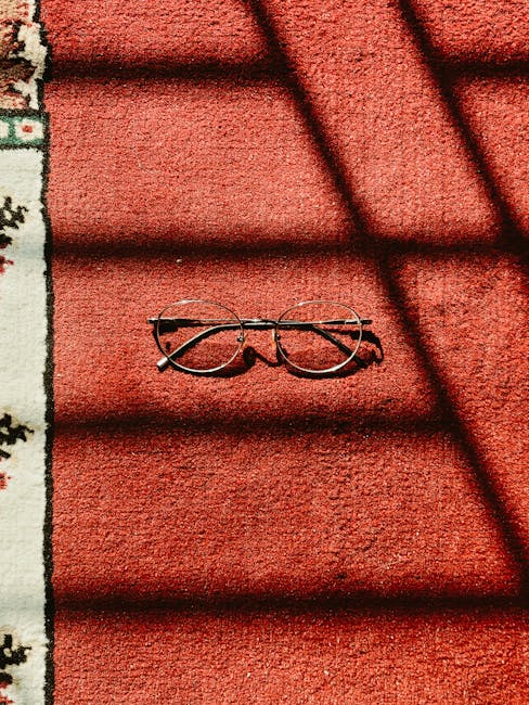 A pair of round eyeglasses with thin metal frames rests on a red outdoor carpet or mat, with shadows from nearby window or door frames cast across the surface. The red textured material appears clean and well-maintained, situated next to a decorative edge of a patterned rug or mat. The lighting highlights the vibrant color and subtle surface details, indicating a bright, sunny day outside. The scene is part of a residential or commercial setting, showcasing a neat and tidy space, consistent with surface cleaning and maintenance services provided by Carpet Cleaning Pimlico in Pimlico SW1V for the Eccleston Square area.