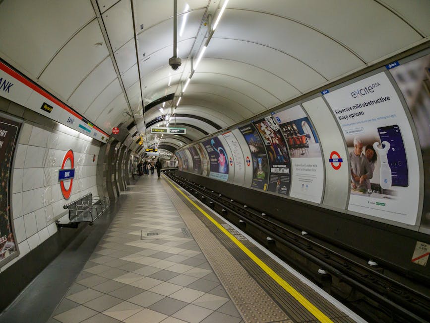 An underground station platform at Pimlico, featuring tiled flooring with a geometric pattern in shades of gray, white, and black. The curved white ceiling has embedded lighting panels illuminating the station. On the left side, there is a tiled wall with an iconic London underground roundel sign and a black metal bench with a perforated seat. The right side has a series of large, colorful advertising posters mounted behind a stainless steel curved wall that encloses the platform edge. The ash-colored tracks run parallel to the platform, separated by a tactile yellow warning strip adjacent to the edge. Several passengers are walking along the platform in the background. This scene is associated with a post-event deep cleaning service provided by Carpet Cleaning Pimlico, focused on maintaining hygiene and cleanliness in busy public transportation environments.