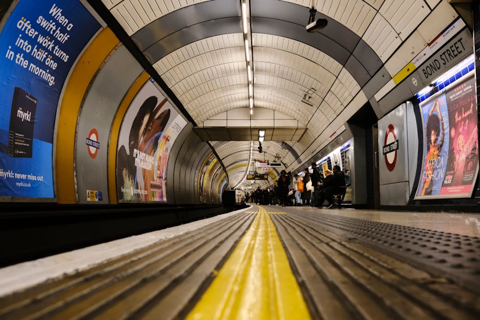 An underground station platform at Pimlico, featuring tiled flooring with a geometric pattern in shades of gray, white, and black. The curved white ceiling has embedded lighting panels illuminating the station. On the left side, there is a tiled wall with an iconic London underground roundel sign and a black metal bench with a perforated seat. The right side has a series of large, colorful advertising posters mounted behind a stainless steel curved wall that encloses the platform edge. The ash-colored tracks run parallel to the platform, separated by a tactile yellow warning strip adjacent to the edge. Several passengers are walking along the platform in the background. This scene is associated with a post-event deep cleaning service provided by Carpet Cleaning Pimlico, focused on maintaining hygiene and cleanliness in busy public transportation environments.