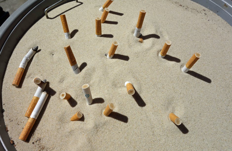 Close-up view of a circular sandpit with a light beige, fine-textured surface in a residential or commercial outdoor setting. Several cigarette butts and filters are scattered across the sand, some with burnt tips and others intact. The surrounding edge of the sandpit is visible, constructed from a dark material, possibly metal or plastic, with a curved shape. Shadows cast by the cigarette butts and filters indicate strong sunlight. The scene reflects an area requiring cleaning or maintenance to remove debris and ensure hygienic, tidy outdoor spaces. This image highlights the importance of surface cleaning and sanitisation in maintaining a clean environment, as promoted by Carpet Cleaning Pimlico.