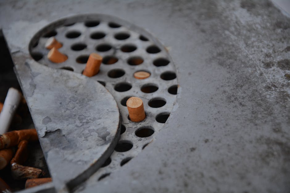 Close-up of a kitchen or bathroom floor with a circular drain cover and surrounding concrete surface. The drain cover is metallic with numerous round holes and has several scattered cigarette butts resting on it and nearby. The concrete surface appears slightly dusty and weathered, with some marks and minor debris. The lighting is neutral, highlighting the rough texture of the floor and the metallic sheen of the drain cover. This image showcases the importance of surface cleaning and sanitisation in maintaining hygiene, as offered by Carpet Cleaning Pimlico, especially when addressing issues like cigarette odour removal from property carpets and flooring.