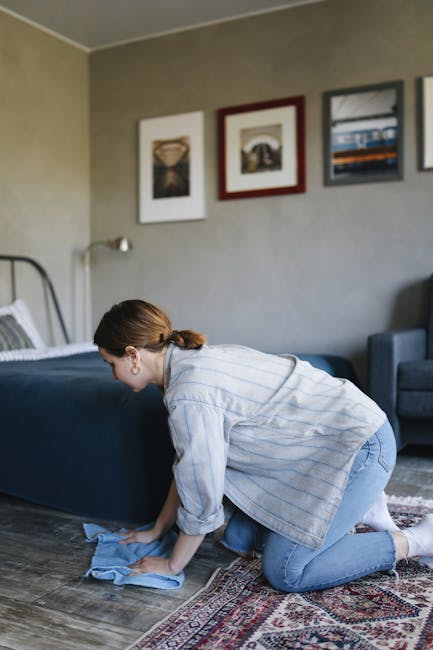 A woman with brown hair tied back, wearing a light blue striped shirt and jeans, is kneeling on a decorative rug while cleaning the wooden floor in a bedroom with a blue bed frame. She is using a blue cloth to wipe the floor. The room has beige walls adorned with three framed art pieces, a grey sofa, and natural light entering from the window, highlighting the clean, polished surface. This scene illustrates domestic cleaning and surface sanitisation, aligning with the services offered by Carpet Cleaning Pimlico for same-day quotes in Pimlico.