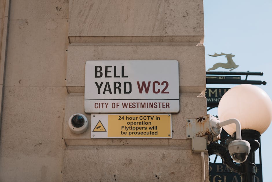 Close-up of a beige stone wall featuring a white and red sign displaying 'BELL YARD WC2' with the text 'CITY OF WESTMINSTER' underneath, and a smaller yellow sign warning about 24-hour CCTV surveillance and prosecution of flytippers, alongside a security camera and a white street lamp with a rounded opaque globe, with a clear blue sky in the background. The area appears clean and well-maintained, with no visible dust or debris, and the lighting suggests daylight conditions. The image captures the urban residential or commercial environment associated with Westminster Council regulations, aligning with surface cleaning and maintenance themes relevant to Pimlico.