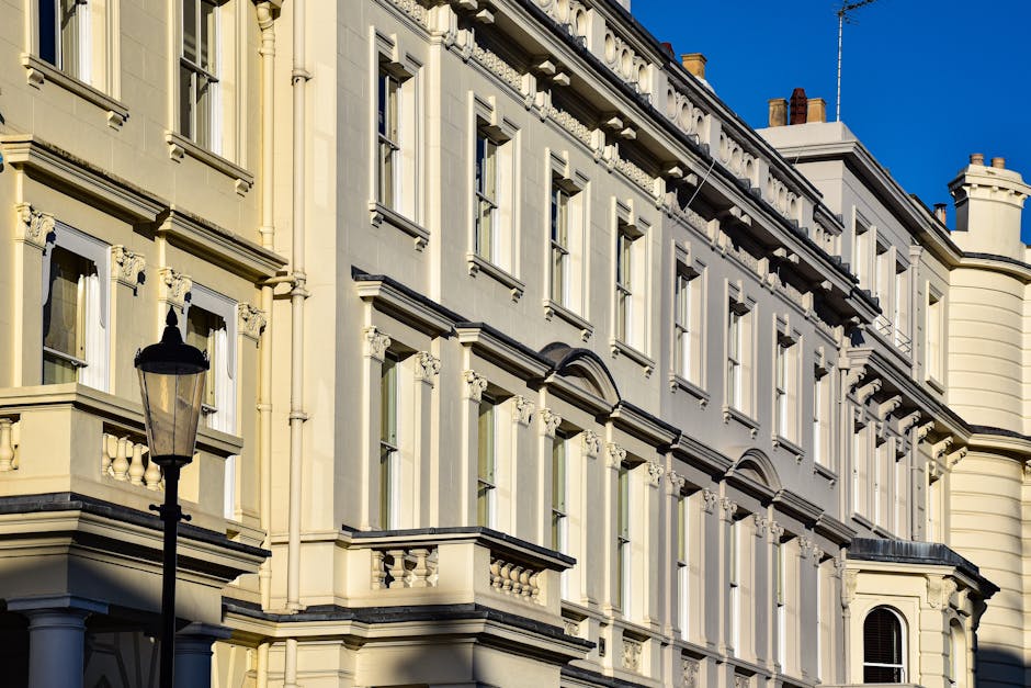 Photograph of a Victorian-style residential building with cream-colored stucco facade, multiple large sash windows framed with decorative molding, and a small balcony with a balustrade. A black street lamp is positioned in front of the building, casting a shadow on the wall. The bright sunlight highlights the clean, well-maintained surface of the building's exterior, emphasizing the architectural details and the tidy appearance. The scene captures the elegant, historic architecture typical of Pimlico, with a clear blue sky in the background. This image visually represents the importance of surface cleaning and maintenance for aesthetic and hygienic purposes as provided by Carpet Cleaning Pimlico, a specialist in domestic cleaning services in the Westminster area.