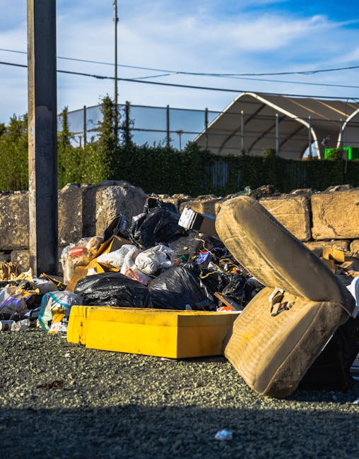 A pile of outdoor waste including black garbage bags, a yellow plastic container, an old, dirty car tire, and scattered litter, situated on gravel near a stone wall with metal fencing in the background. The scene is illuminated by natural daylight, with fencing and some greenery visible beyond the wall, indicating an outdoor setting possibly in Pimlico. The image highlights issues of rubbish accumulation and cleanliness management, relevant to property maintenance and hygiene practices in the area, as addressed by Carpet Cleaning Pimlico in the context of Westminster Council rules for mattress disposal and urban cleanliness.