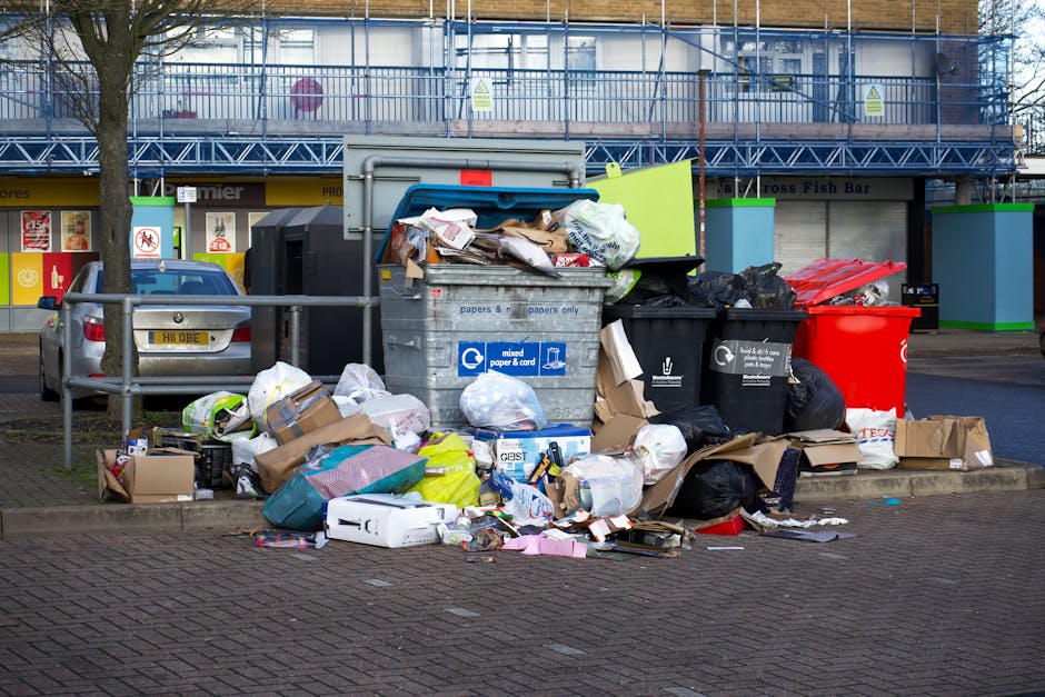 A pile of outdoor waste including black garbage bags, a yellow plastic container, an old, dirty car tire, and scattered litter, situated on gravel near a stone wall with metal fencing in the background. The scene is illuminated by natural daylight, with fencing and some greenery visible beyond the wall, indicating an outdoor setting possibly in Pimlico. The image highlights issues of rubbish accumulation and cleanliness management, relevant to property maintenance and hygiene practices in the area, as addressed by Carpet Cleaning Pimlico in the context of Westminster Council rules for mattress disposal and urban cleanliness.